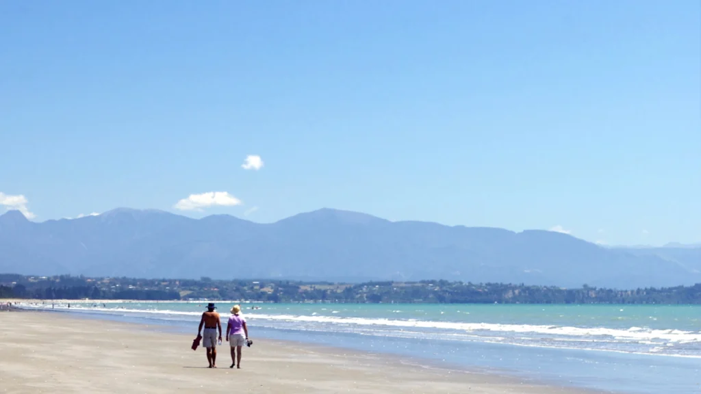A couple strolling along a sunny beach in NZ reflecting on the choice to transfer superannuation to KiwiSaver, a key financial planning move